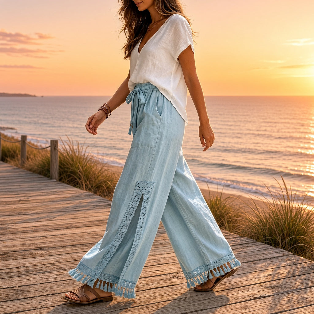 Nahar Bloom – Calças Fluidas de Perna Larga com Racha e Franjas, cor Azul-Claro, mulher a caminhar num passadiço de madeira junto ao mar ao pôr do sol